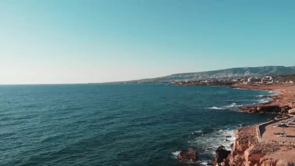 Scogliere rocciose colpite da acqua cristallina blu oceano schizzi e creando schiuma bianca con montagne all'orizzonte e alberghi e resort di baia di corallo. ciprus riva del mare con scogliere rocciose e bordi — Video Stock