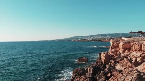 Drone vuela sobre acantilados y playa rocosa en ciprus con montañas en el horizonte y olas del océano golpeando la costa rocosa y la playa. Vista aérea de acantilados rocosos de playa y fuertes olas marinas que golpean la costa — Vídeos de Stock
