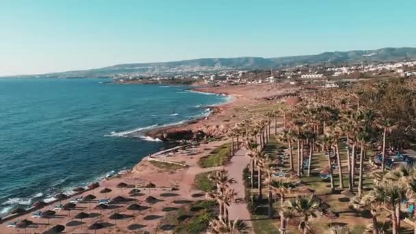 Drone voa sobre a área do hotel com pinheiros e palmas. Ondas do mar atingindo costa rochosa com topos de montanha no fundo. Vista aérea do passeio marítimo com hotéis, palmas e caminho para pedestres — Vídeo de Stock