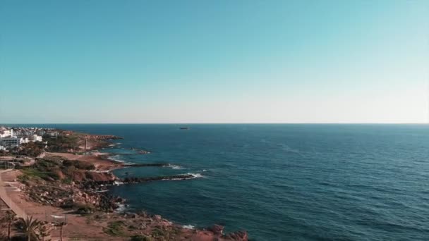 Вид на блакитне море з маленьким покинутим човном на горизонті. Сторм'яні морські хвилі, що вражають кам'янистий пляж, створюють білу піну і розбризкують. Вид з повітря на готелі та курорти в бухті Корал Кіпр — стокове відео