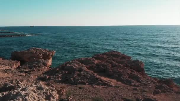 Mavi okyanus suyu ve ufukta güneş olan kahverengi kayalıklar. Kıbrıs'ta mercan plajı. Kayalıklar ve kayalar ile çarpışan mavi okyanus suyunun havadan görünümü. — Stok video