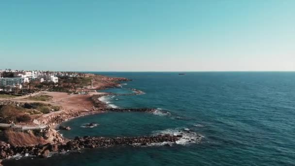 Letecký výhled na skalnaté pobřeží na Kypru s hotely a mořským poloostrovem. Drone na ostrovní skalnaté pláži a bouřné vlny se střetává se útesy a skalami. Opuštěný člun na obzoru v čisté vodě — Stock video