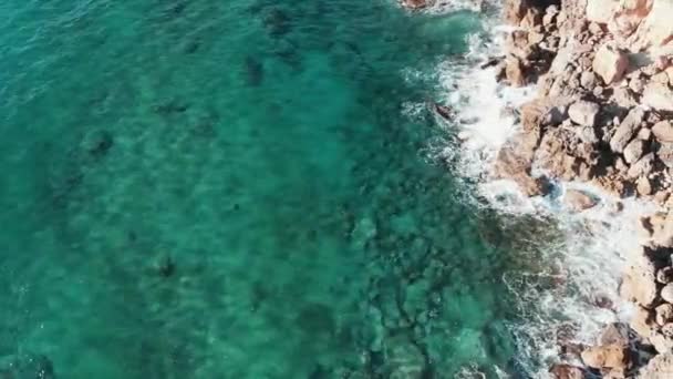 Drone flyger över transparent blått klart havsvatten med vågor som kollapsar mot klippiga bergsklippor som plaska och skapar vitt skum. Top antenn skott av havsvågor och klippor — Stockvideo