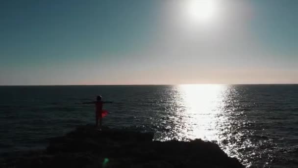 Mavi okyanusta güneş ve güneş yoluna karşı elleri havada kayalıklarda duran kadın silueti. Güçlü rüzgarda çırpınan kırmızı elbiseli kayalık kayalıklarda duran kolları dışında kız Drone görünümü — Stok video