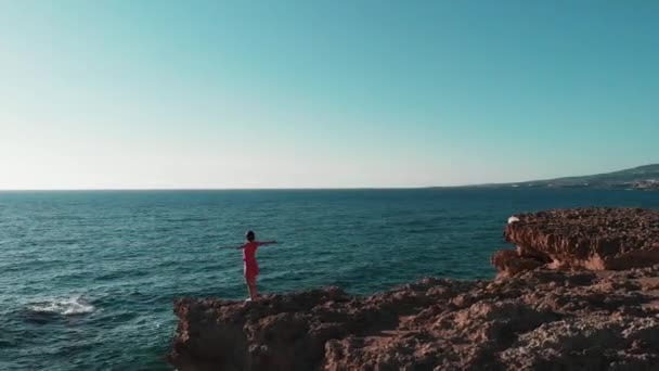 赤いドレスを着て岩の崖の上に立つ若い女性は、空中で手を離して、風に羽ばたくドレス。新しい海を離れて腕を持つ岩の崖の上で暖かい風と太陽を楽しむ魅力的な女性 — ストック動画