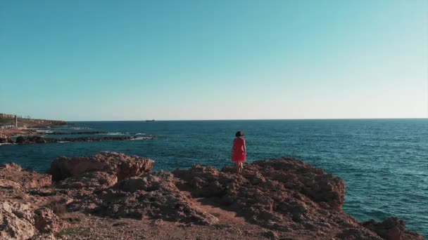 Bella ragazza che cammina su grandi rocce. Donna caucasica in piedi sulla spiaggia rocciosa. La signora in abito rosso alza le braccia ai lati. Ragazza in abito corto guardando l'orizzonte. Bruna donna in piedi su grandi scogliere — Video Stock