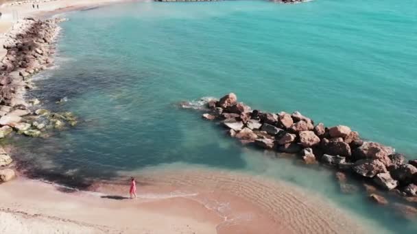 Girl on beach. Young woman in red dress walking along rocky stones. Attractive girl on sandy beach. Cute caucasian lady in red dress. Aerial view of sandy beach with clear sea water. Sea landscape — Stock Video