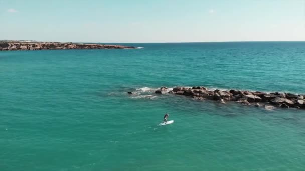 Drone flies near beautiful rocky seashore. Aerial view of young sportive man on sup. Attractive boy floating along rocky stones. Ocean with blue clear water and rocks. Drone shot of sea landscape — Stock Video