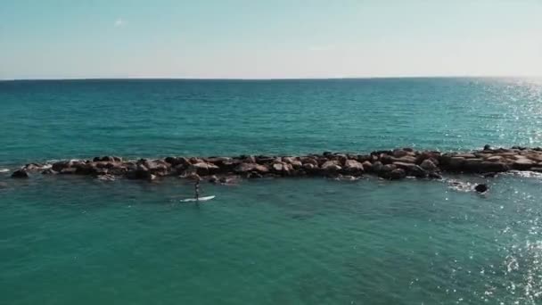 Ember lebeg a sup. Fiatal fiú szörfözés. Légi kilátás a gyönyörű tengeri tájra. Drone lövés a tengeri sziklás kövek. Kék tiszta vizű tengeren úsznak. Férfiak relaxáló és élvezi a nap — Stock videók