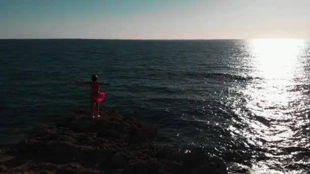 岩石海滩上的女孩站在海洋悬崖上的年轻女子女孩在海上穿红色衣服。迷人的女人看着夕阳。美丽的海洋日落。站在大岩石石上的女孩的空中无人机视图 — 图库视频影像