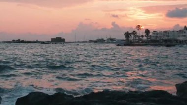 Baf şehrinde güneş, kayalık plaja çarpan dalgalarla batıyor ve köpük yaratıyor. Kale ve palmiye ağaçları ile şehir gezinti ve rıhtım ile arka planda marina. Yavaş çekim
