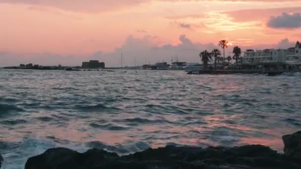 嵐の海と夕日は、パフォスキプロスの岩の海岸線をヒット.ヤシの木を背景に城と夜の遊歩道とマリーナ岸壁。旅行コンセプト — ストック動画