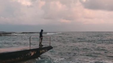 Fırtınalı okyanusa bakan yalnız çocuğun yan görünümü. Ufka bakan genç üzgün çocuk. Melankolik çocuk iskelede duruyor. Issız sahilde genç yalnız adam