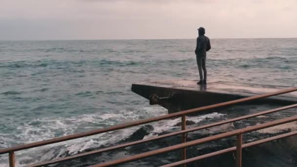 Un ragazzino malinconico in piedi al molo. Ragazzo solitario che guarda il mare tempestoso e piovoso. Uomo pessimista al porto turistico. Giovane ragazzo in abito scuro in piedi in riva al mare — Video Stock