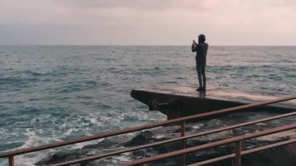 Ung pojke tar bilder av stormiga havet. Man gör foto stående vid piren. Lonely Boy tar video av blåsigt hav. Side View of Young man tar bilder på Seacoast — Stockvideo