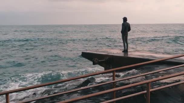 Un ragazzino malinconico in piedi al molo. Ragazzo solitario che guarda il mare tempestoso e piovoso. Uomo pessimista al porto turistico. Ragazzo vestito di scuro, in piedi sulla costa. Grandi onde oceaniche colpiscono rocce. Rallentatore — Video Stock