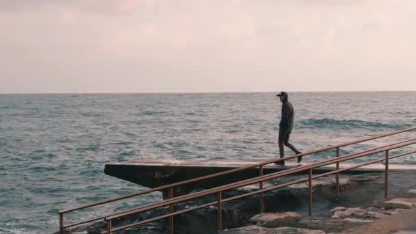 年轻的孤独的男孩走在长廊。悲伤的人站在码头上。忧郁的男孩看着暴风雨的海洋。孤独的男孩来到海边。慢动作 — 图库视频影像