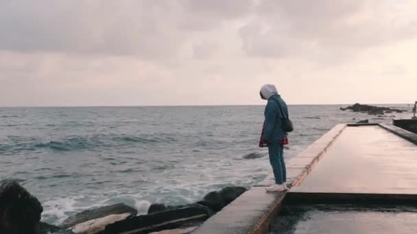Young lonely girl standing at pier at rain. Lonesome girl looking at stormy sea. Woman stands at deserted promenade. Sad beautiful girl looking at ocean. Side view of lonely girl near sea — Stock Video