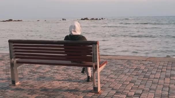 ビーチの近くのベンチに座ってフード付きの黒いジャケットを着た若い女性。嵐の海を見ている女性。雨天のベンチに座っている女の子。木製のベンチに座っている若い孤独な少女の裏側図 — ストック動画