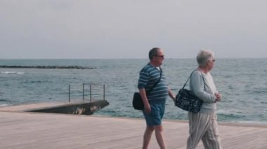 16 Mart 2019/Kıbrıs, Baf. Deniz plajı boyunca yürüyen insanların olduğu turist iskelesi. Yaşlı hoş çift gezinti boyunca geziniyor. Mutlu bir aile iskelede yürüyor. Akdeniz'in güzel manzarası