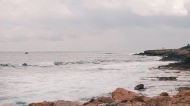 Akdeniz'in güzel büyük kayalık plaj. Büyük okyanus dalgaları kayalık deniz kıyılarını vuruyor. Okyanus dalgaları köpük ve sıçrama yaratıyor. Açık denizde yelken küçük balıkçı teknesi. Denizde beyaz küçük gemi