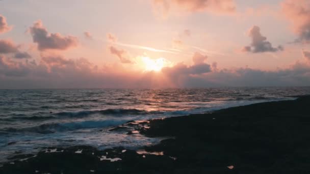 夕日が雲の後ろに隠れている。海辺の美しいピンクの夕日。岩の多い海岸の美しい日没。ピンクの夕日と岩のビーチでロマンチックな夜。ピンクの空に対する岩のビーチのシルエット. — ストック動画