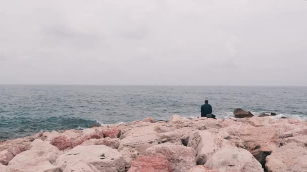 Lonely smutny człowiek siedzi na wielkim kamieniu w pobliżu oceanu. Pracownik biurowy siedzi w pobliżu morza po dniu roboczym. Odosobniony samotny chłopiec patrząc na morze. Rocky Seacoast z dużymi falami. Morze Śródziemne z dużymi falami — Wideo stockowe