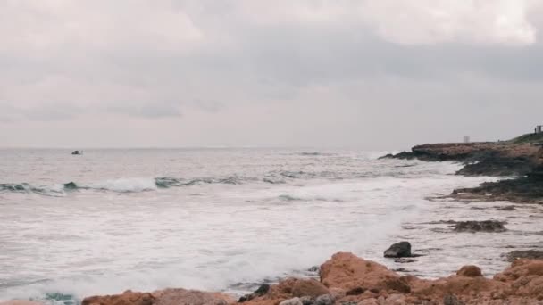 地中海の美しい大きな岩のビーチ。岩の多い海岸を襲う大きな海の波。泡と水しぶきを作成する海の波。オープンウォーターで航行する小さな漁船。スローモーション — ストック動画