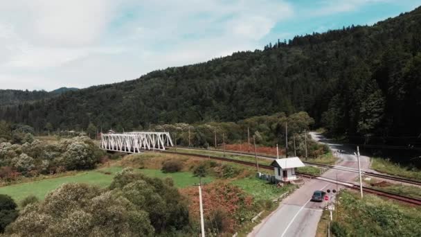 Dron leci wzdłuż linii kolejowej w Karpatach. Lotnicze Drone widok gór z mostu kolejowego przeszedł przez rzekę. Przejazd kolejowy w górach. Przejazd samochodem przez przejście kolejowe — Wideo stockowe