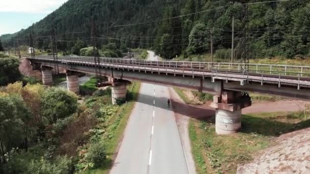 Drönare flyger i bergslandskapet med gröna träd och flod. Järnvägsbron från fågelperspektiv. Top utsikt över gamla betongbro. Kvinna cyklist utbildning på cykel i bergen. Flicka cykling på väg — Stockvideo