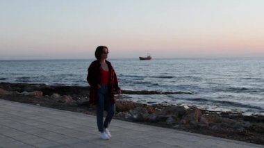 Siyah güneş gözlüğü genç esmer kadın gün batımında gezinti üzerinde yürüyor. Rahat giyim çekici kız plaj boyunca yürüyüş ve okyanus pembe günbatımı bakarak. Eski gemi denizde mahsur kaldı. Yavaş çekim