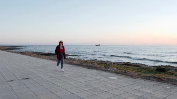 Kaukasische brunette meisje in zonnebril, rood shirt en jeans wandelen langs het strand. Jonge charmante vrouw in rood badpak en blauwe jeans loopt langs de strandpromenade. Slow Motion — Stockvideo