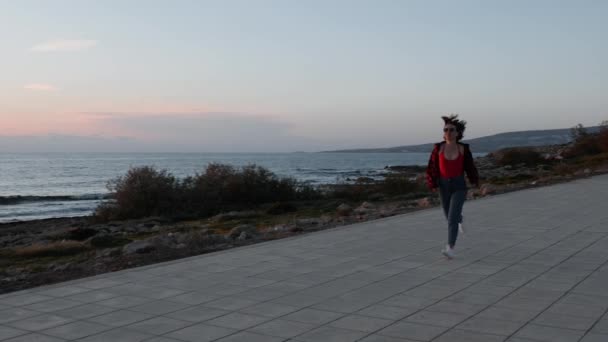 Jonge hete meid in rood badpak en blauwe jeans loopt langs het strand. Kaukasische brunette vrouw in rood shirt en jeans lopen op de promenade bij zonsondergang. Jong gelukkig meisje loopt bij zonsondergang. Slow Motion — Stockvideo