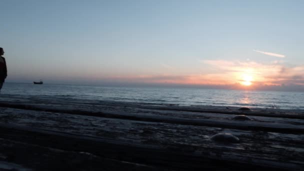 Młoda samotna dziewczyna w dorywczo nosić chodzenie wzdłuż skalistej plaży ze statkiem na tle. Kaukaski brunetka kobieta chodzenie na promenadzie. Sylwetka młodej dziewczyny chodzącego na nabrzeżu o zachodzie słońca — Wideo stockowe