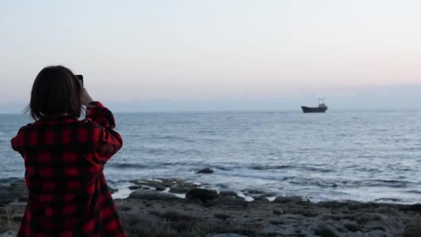 Chica morena atractiva tomando fotos de la vieja nave. Chica joven en camisa de cuadros rojos tomando fotos de mar al atardecer. Mujer tomando fotos del océano en el teléfono inteligente . — Vídeo de stock