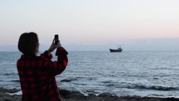 Молодая кавказская девушка фотографирует старый корабль, застрявший в море. Привлекательная девушка в красной клетчатой рубашке фотографирует морской пейзаж. Женщина фотографирует океан и корабль. Старый корабль, застрявший в море . — стоковое видео