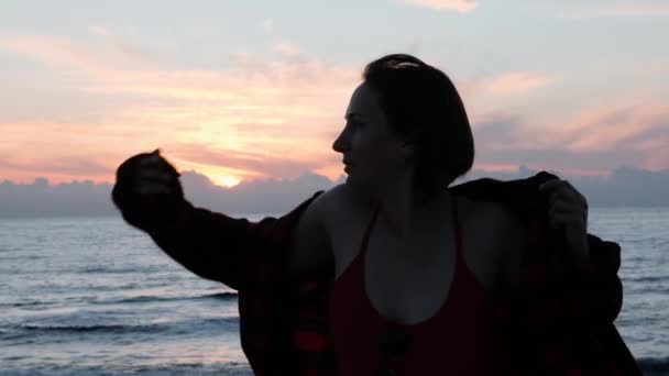高加索少女穿上红色衬衫对抗大海和日落。迷人的黑发女人在黄昏时分摆姿势对海。迷人的女孩微笑的相机。美丽的年轻女子穿上衬衫在海边 — 图库视频影像