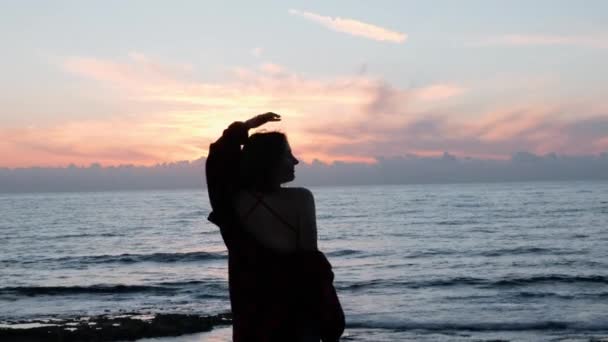 日没時にカメラのためにポーズホットブルネットの女の子。岩の多い海岸で笑顔と回転する若い女性のシルエット。夕暮れ時に海岸でカメラのポーズをとる赤い水着とジーンズの魅力的な女の子. — ストック動画