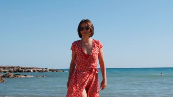 Close up side view of young brunette girl on sandy beach. Close up of attractive hot girl walking on sea shore. Close up of caucasian girl in red short dress and black sunglass. — Stock Video