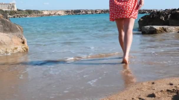 Young caucasian girl comes into water at coast of sea. Brunette girl in short red dress walking on seashore. Attractive woman walking through sea shore. Young attractive girl at sea. Slow motion — Stock Video