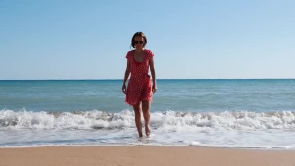 Kaukasische Brünette in rotem Kleid und schwarzer Sonnenbrille am Sandstrand. attraktive Frau geht vom Meer aus vor die Kamera. Mädchen kommt mit azurblauem Wasser aus dem Meer. junges heißes Mädchen geht am Strand — Stockvideo