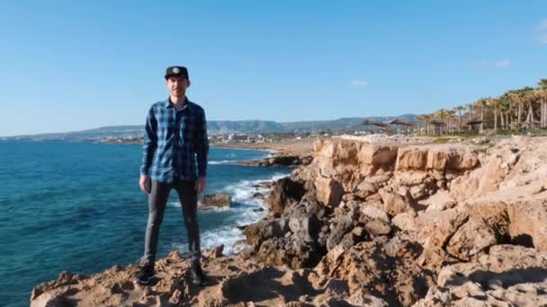 Front view of young man standing on edge of cliff and doing dab pose. Man with dab pose. Attractive man standing on cliff and smiling. Young guy standing on rocks surrounded by sea and fooling around — Stock Video