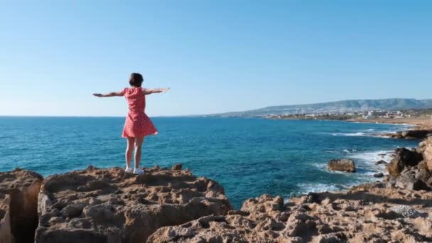 Atrakcyjna dziewczyna stoi na skraju urwiska z wyciągniętymi ramionami do boków. Brunetka kaukaski dziewczyna w czerwonej sukni stoi na blef i patrząc na morze. Widok z tyłu dziewczyny stoi na klifie w pobliżu morza — Wideo stockowe