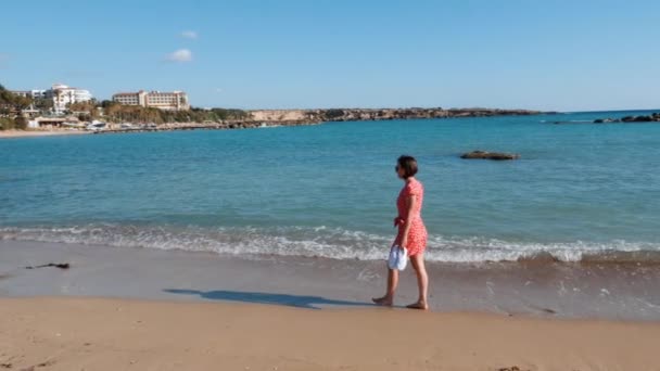 Молодая очаровательная одинокая девушка прогуливается вдоль песчаного пляжа у моря. Привлекательная кавказка в красном платье гуляет по морскому пляжу. Босоногая девушка гуляет по песчаному пляжу. Одинокая девушка ходит по морю. Медленное движение — стоковое видео