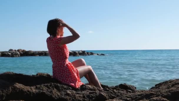 Junges schlankes kaukasisches Mädchen in rotem kurzen Kleid, das auf einem großen Felsen am Meer sitzt. attraktive brünette Frau glättet die Haare sitzt in der Nähe felsigen Strandes. charmante heiße Mädchen sitzt auf einem Felsen in der Nähe des Meeres und sonnt sich — Stockvideo