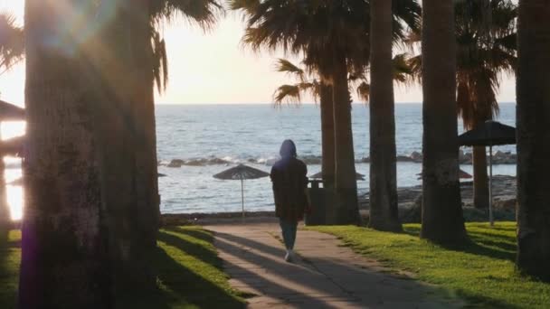 Junges Mädchen in Freizeitkleidung geht an den Strand. brünette Frau zu Fuß zum Strand. Mädchen, das mit Palmen durch die Touristengegend läuft. Frau geht auf Hotelgelände mit Palmen und Sonnenschirmen. Zeitlupe — Stockvideo