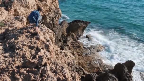 O caucasiano atraente senta-se na rocha à beira do penhasco. O rapaz desce ao penhasco e olha para o mar. O tipo senta-se na rocha rodeado de mar. Homem senta-se na borda do penhasco — Vídeo de Stock