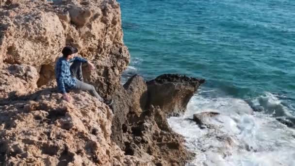 Νέος άντρας με μπλε πουκάμισο και καπέλο να κάθεται πάνω σε βράχια και να κοιτάζει τη θάλασσα. Ένας τύπος που καθόταν στην άκρη του γκρεμού. Ο αρσενικός τουρίστας αναπαύεται σε βράχους και απολαμβάνει τη θάλασσα. Αγόρι που ποζάρει στην άκρη του γκρεμού. Τύπος που κοιτάζει τα κύματα της θάλασσας — Αρχείο Βίντεο