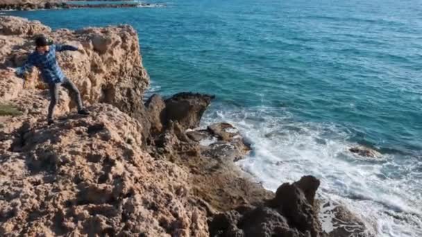 Un joven se para en las rocas y arroja piedras al agua. Niño caucásico arrojando piedras al mar de pie en el borde del acantilado. Hombre de pie sobre rocas rodeadas de mar. Solitario parado en el borde del acantilado — Vídeos de Stock