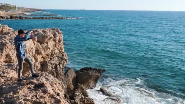 Um tipo a atirar pedras à água. O jovem está à beira do penhasco e atira pedras ao mar. Menino de camisa azul e boné em pé sobre rochas e joga pedras na borda do penhasco. Movimento lento — Vídeo de Stock